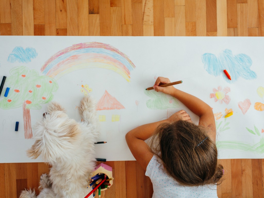 Little Girl lying on the floor and drawing with crayons on a paper, together with her dog.