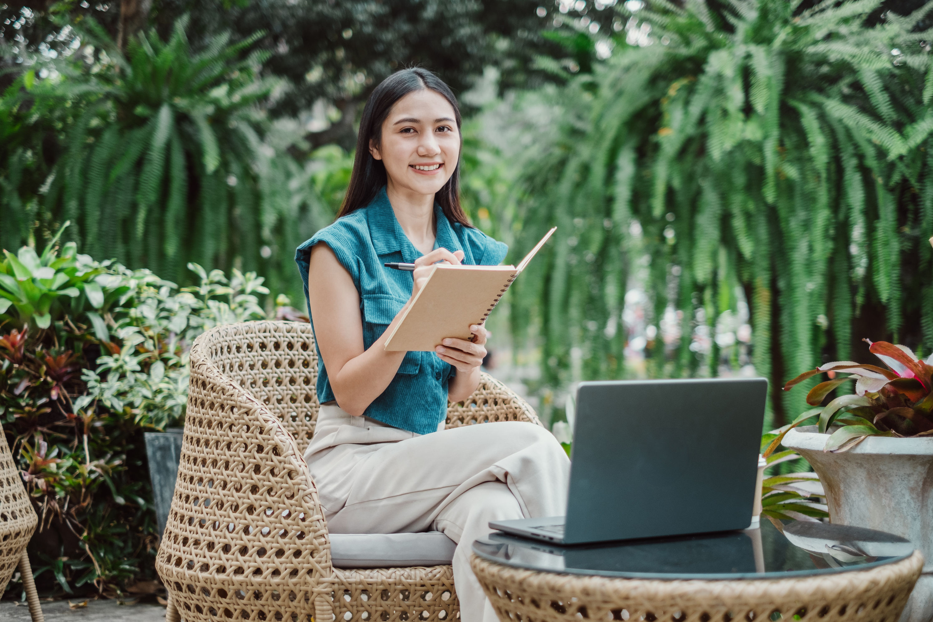 A lady sitting with a notebook and a laptop surrounded by greenery.